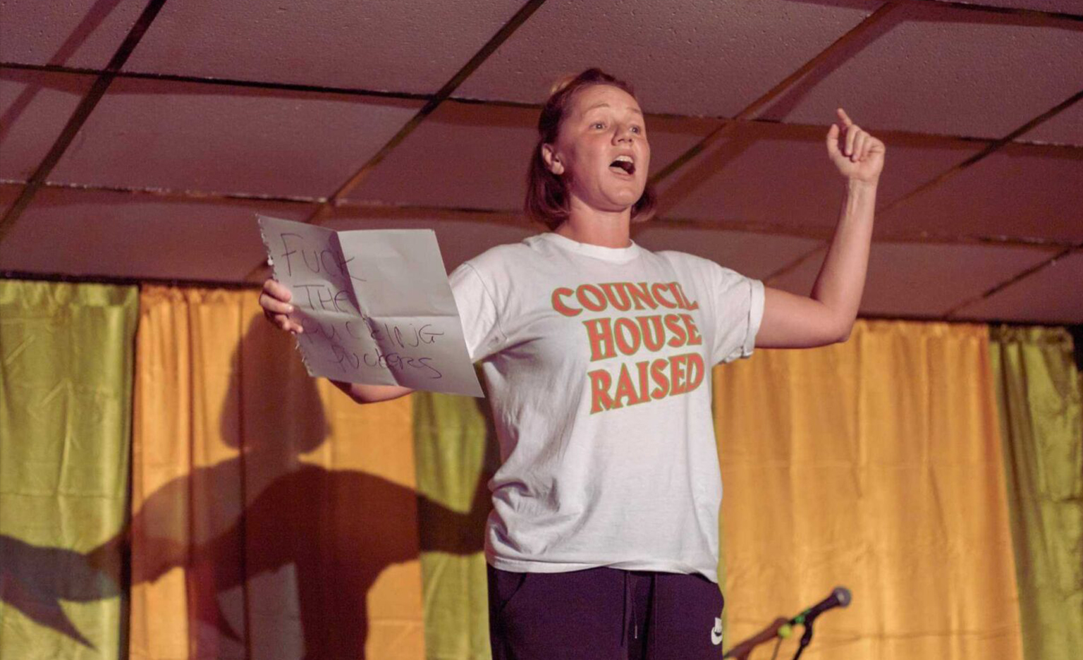 Jodie rehearsing. She is standing in a low ceiling room, yellow and pistachio green curtains behind her. She wears a white t-shirt with all caps orange letters on it that read: "Council house raised". She holds a white sheet of paper with one hand, on which can be seen the words: "FUCK THE FUCKING FUCKERS". Her other hand is raised up in the air in protest and her faces suggests shouting.