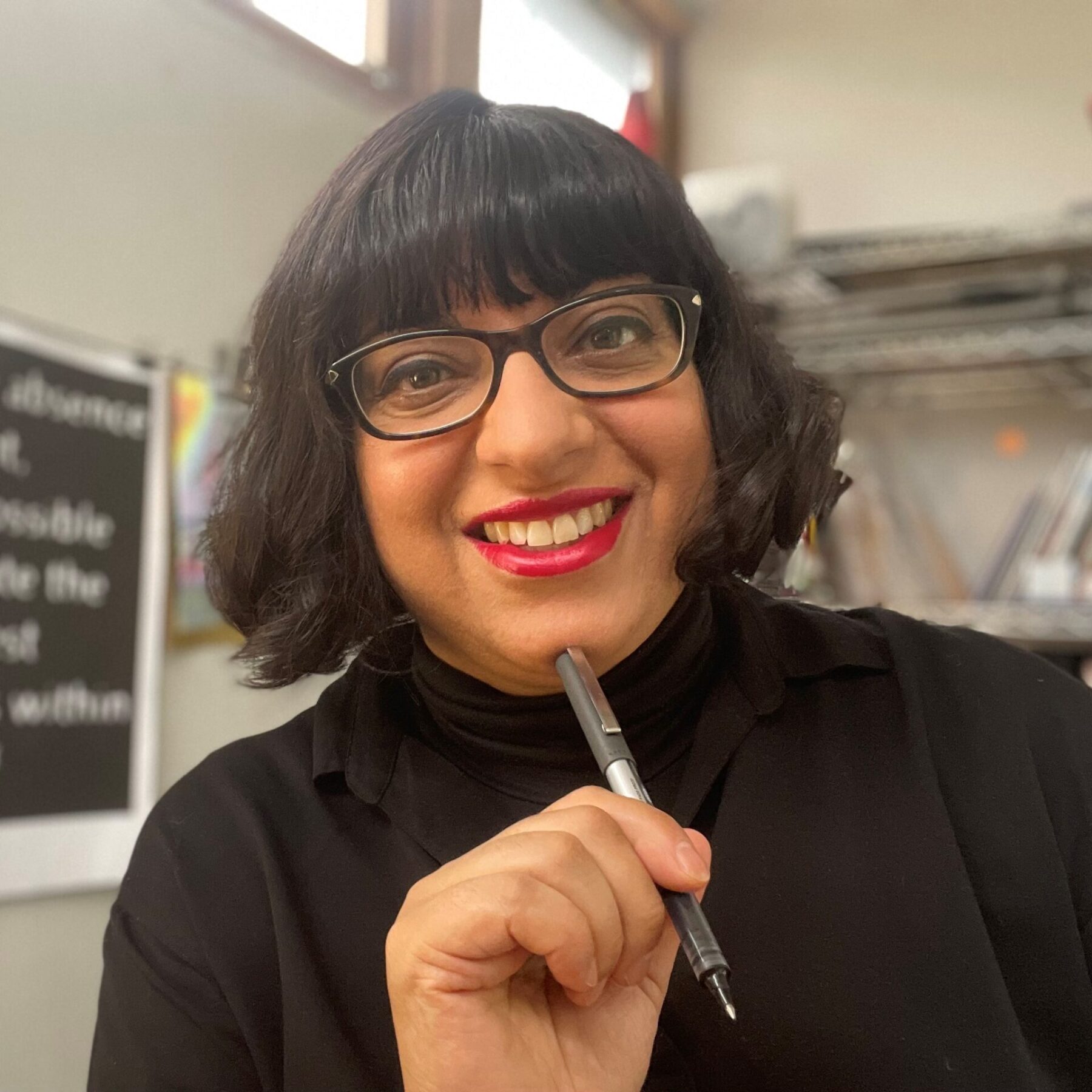 Binita is a middle-aged Indian woman with bobbed black hair and a fringe. She wears brown rectangular glasses, red lipstick and wears black a lot.