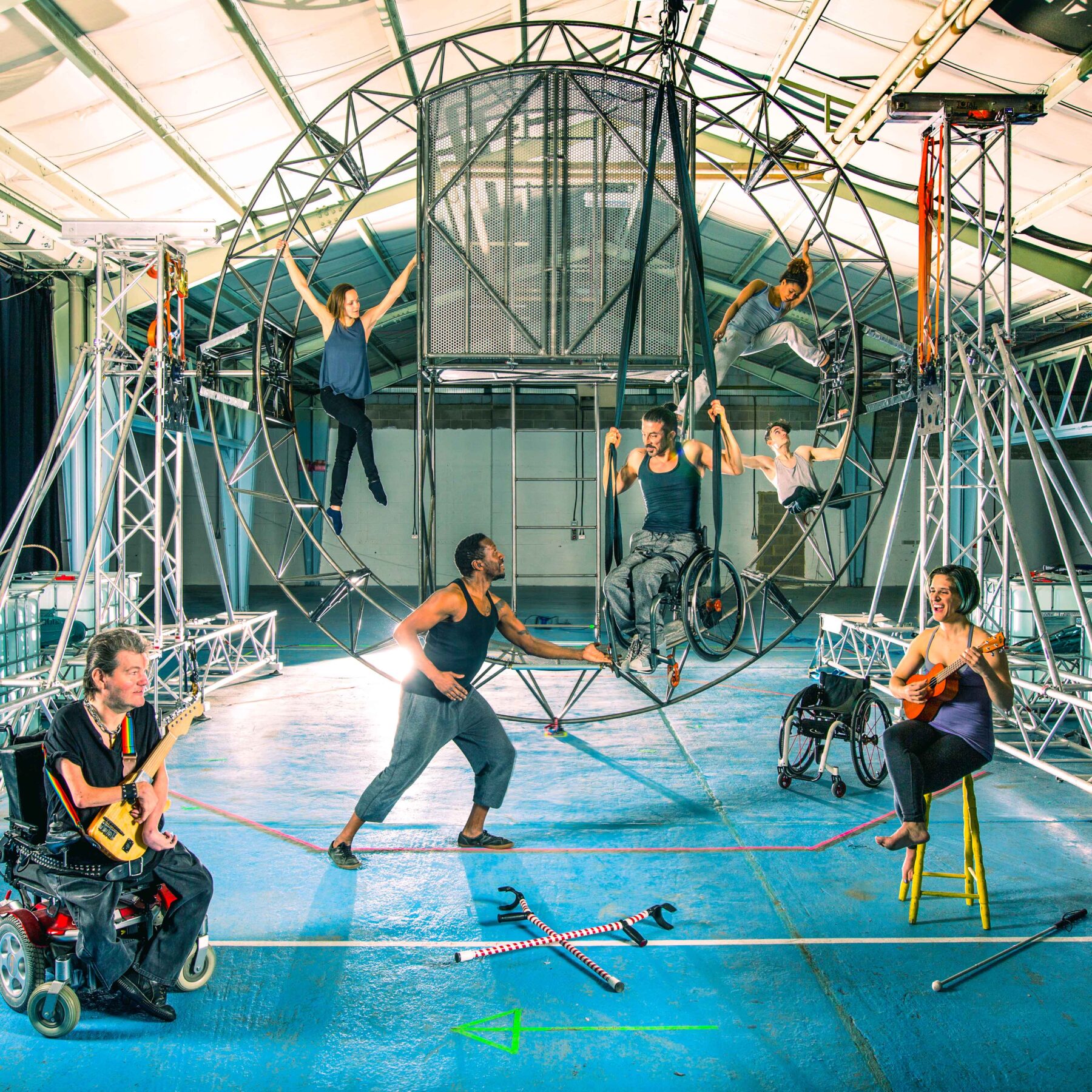 A rehearsal room with a giant steel wheel in the background. There are three performers on it: a white woman hanging on by her arms, a white man who uses a wheelchair held up in the air by some black straps, a young white man with thin legs folded below his body who also holds himself up with his arms and above him a mixed-race woman with one leg straight and the other bent and pressing on the outer side of the steel wheel. In the foreground, a Black man holds onto the wheel while standing on the floor and looks at the performers on the wheel. A white woman on the right sits on a stool and plays the ukulele. On the left, a white man in an electric wheelchair holds a custom-made guitar that fits on his lap.