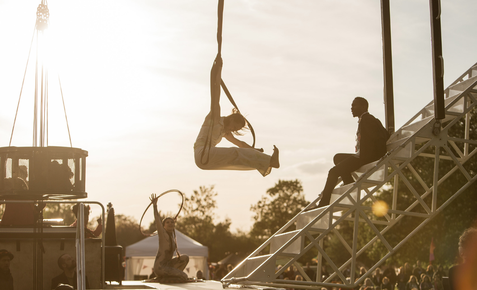 Outdoors settings. The sun burns behind diffuse clouds, making the light soft and warm. Three performers on a stage. The first on is a Black man sitting on metal white stairs. The second one is a white woman sitting with one arm in the air while twirling a hula hoop around a wrist. The third one is a white woman up in the air, holding herself up by holding onto circus straps with one hand. She is bent forward with her legs parallel to the ground.