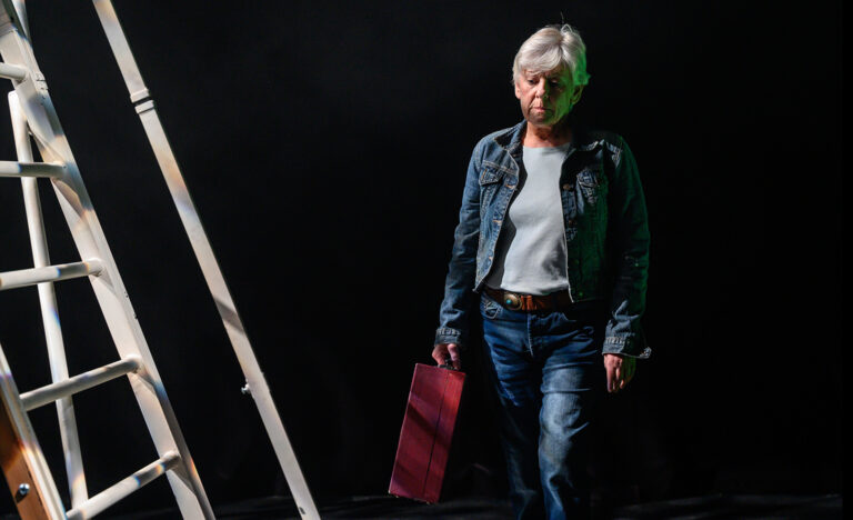 A white woman in her sixties is walking towards the camera, next to a white ladder looking forlornly at the floor. She has short white hair, blue eyes and wears a denim jacket, white shirt, jeans and a brown belt. She is holding a red briefcase.