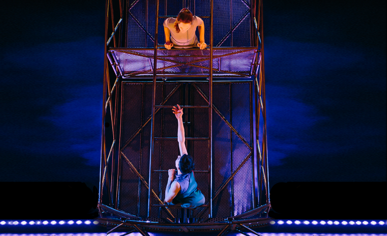 Indoor setting, dark purple lighting. A giant steel structure with two performers on it. The structure looks like a very large wheel, about 3.5 metres high. The first performer is a white man in his twenties. His thin legs are folded below his body and he holds onto the wheel with his arms. We see his back. He is at the bottom of the wheel, reaching one arm upward towards the second performer. The second performer is a white woman. She is at the top of the wheel, looking down at the first performer, lying on her stomach and holding onto the edge of the wheel with both hands.