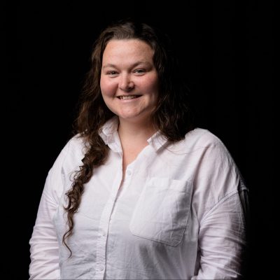 A white woman in er thirties with long brown wavy hair. She wears a white shirt and smiles brightly at us.