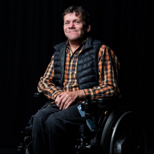 A white man in his forties with short brown hair. He wears a checked orange and yellow shirt and a black sleeveless puffer jacket. He is sitting in his wheelchair.