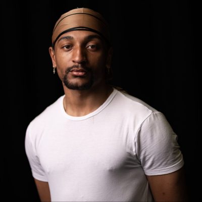 A young Black man with a goatee beard and a silk light brown bandana. He wears a white T-shirt and and one dangly earring on his left ear.