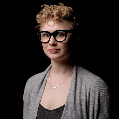 A white woman in her thirties with short curly blonde hair and thick black glasses. She wears a grey cardigan and a black top.