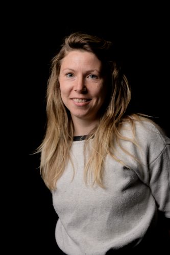 A white woman with long blonde hair flipped to the side. She is wearing a grey shirt and smiling at us.
