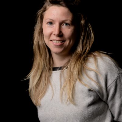 A white woman with long blonde hair flipped to the side. She is wearing a grey shirt and smiling at us.