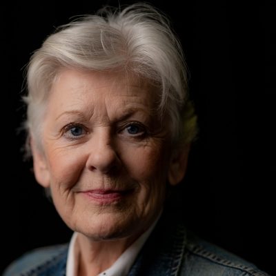 A white woman in her sixties. She has short white hair and blue eyes and wears a denim jacket.