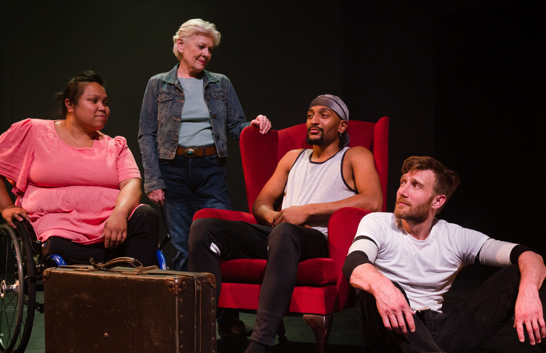 Four performers looking fondly at each other in a dimly lit indoor studio space. In order from left to right is a woman wearing a bright pink shirt who is in a wheelchair, a white woman in her sixties with white hair wearing a denim jacket, a young black man with a goatee sat in a red armchair and a white man with ginger hair and beard sat on the floor.