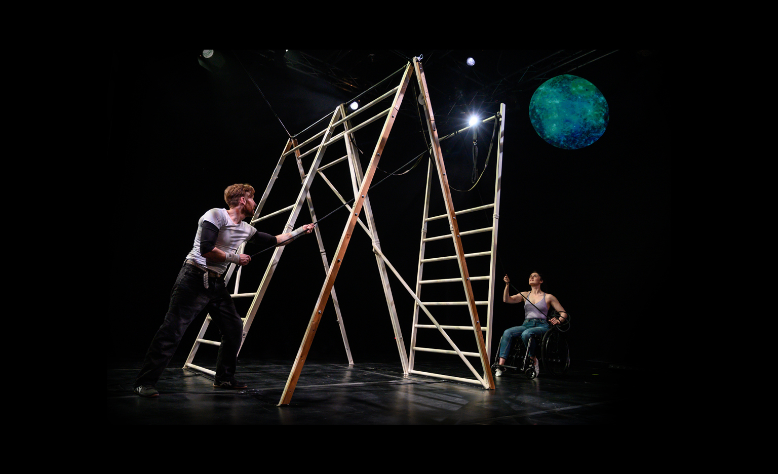 A dimly lit indoor studio. There are two performers standing on opposite ends of a makeshift structure that looks like multiple ladders put together to become one ladder. There is a projection of the earth on the top right hand side. The first performer is a white man with ginger hair who is wearing a white t-shirt and black trousers. The second performer a white female who is a wheelchair user, wearing a purple tank top and blue jeans.